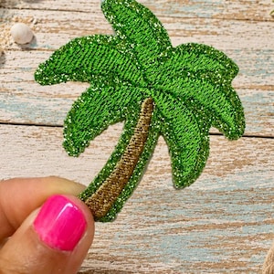 May include: A palm tree patch featuring vibrant green embroidered leaves with a subtle glittery finish and a textured brown trunk. The design is rendered in dense thread work, showcasing a clear silhouette of a tropical tree with detailed stitching.