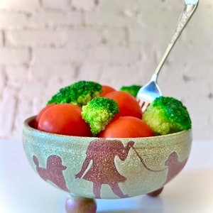 May include: A ceramic bowl with a light green and brown design, featuring a person walking a dog. The bowl is filled with red tomatoes and green broccoli, with a silver fork. The bowl has three small feet.