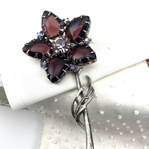 May include: A vintage flower brooch with a silver-tone stem. The brooch features five dark purple, petal-shaped stones surrounding a central clear stone. Small iridescent crystals accent the design, creating a sparkling effect. A classic piece of jewelry.