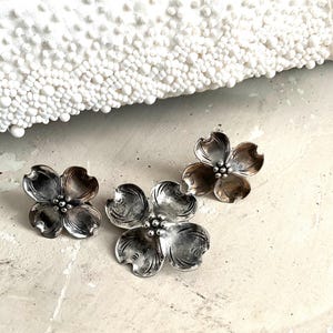 May include: Three silver-toned flower-shaped earrings. The earrings have a detailed design with four petals and a small, round center. The earrings are displayed on a light-colored surface, with a white beaded clutch in the background.