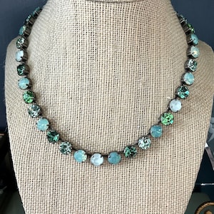 May include: A necklace with a dark metal chain and a series of round, faceted gemstones in shades of green and blue. The necklace is displayed on a mannequin head with a burlap-textured surface, showcasing the jewelry's design and color variations.