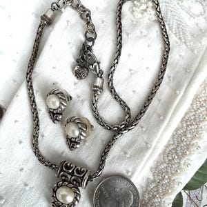 May include: A silver-toned jewelry set featuring a necklace and matching earrings. The necklace has a pendant with an oval pearl, and the earrings also have pearl accents. The set is displayed on a white surface with a quarter for size comparison.
