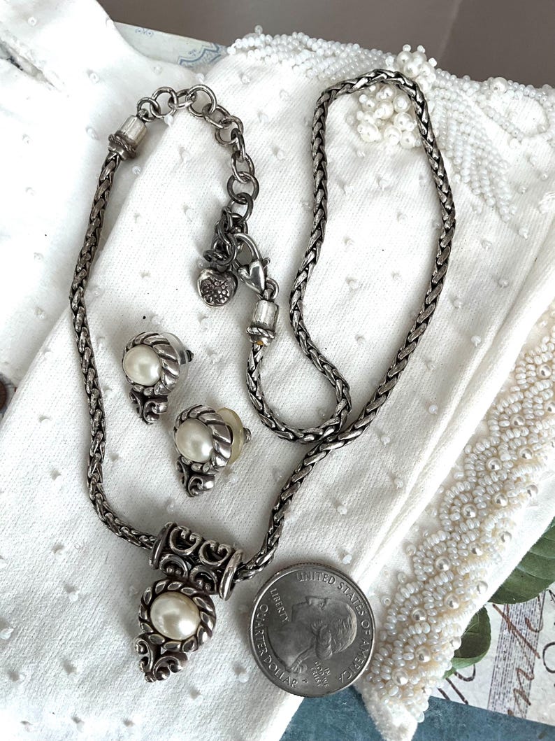 May include: A silver-toned jewelry set featuring a necklace and matching earrings. The necklace has a pendant with an oval pearl, and the earrings also have pearl accents. The set is displayed on a white surface with a quarter for size comparison.
