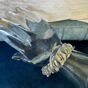 May include: A silver-toned metal bracelet featuring a repeating textured leaf or feather pattern. The bracelet is composed of multiple articulated segments that wrap around the wrist, showcasing a metallic finish with fine etched detailing.