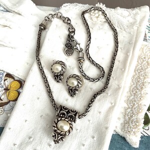 May include: A silver-toned necklace and matching earrings set featuring faux pearls. The necklace has a decorative pendant with an oval pearl, and the earrings are round with a pearl center. The jewelry is displayed on a white surface.