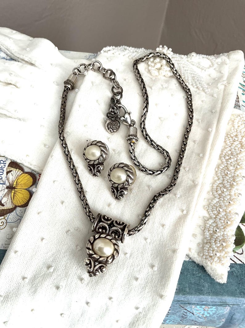 May include: A silver-toned necklace and matching earrings set featuring faux pearls. The necklace has a decorative pendant with an oval pearl, and the earrings are round with a pearl center. The jewelry is displayed on a white surface.