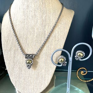 May include: A silver-toned necklace and matching earrings set. The necklace features a woven chain and a pendant with a gold-colored center stone. The earrings have a similar design, displayed on a silver stand. The jewelry is displayed on a beige mannequin.
