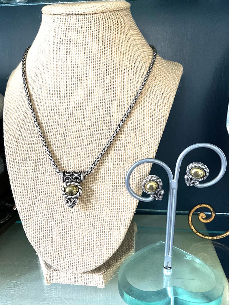 May include: A silver-toned necklace and matching earrings set. The necklace features a woven chain and a pendant with a gold-colored center stone. The earrings have a similar design, displayed on a silver stand. The jewelry is displayed on a beige mannequin.