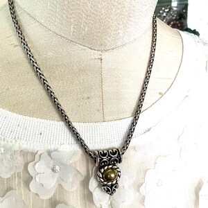 May include: Silver-toned necklace with a braided chain and a decorative pendant. The pendant features a gold-colored orb set within a silver filigree design. The necklace is displayed on a white floral top.