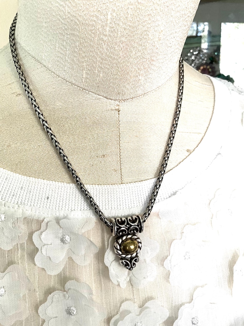 May include: Silver-toned necklace with a braided chain and a decorative pendant. The pendant features a gold-colored orb set within a silver filigree design. The necklace is displayed on a white floral top.