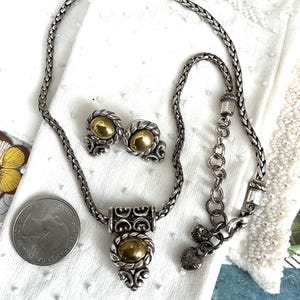 May include: A silver-toned necklace and matching earrings set with gold-colored accents. The necklace features a braided chain and a pendant with an oval gold center. The earrings have a similar design. A US quarter is next to the jewelry.