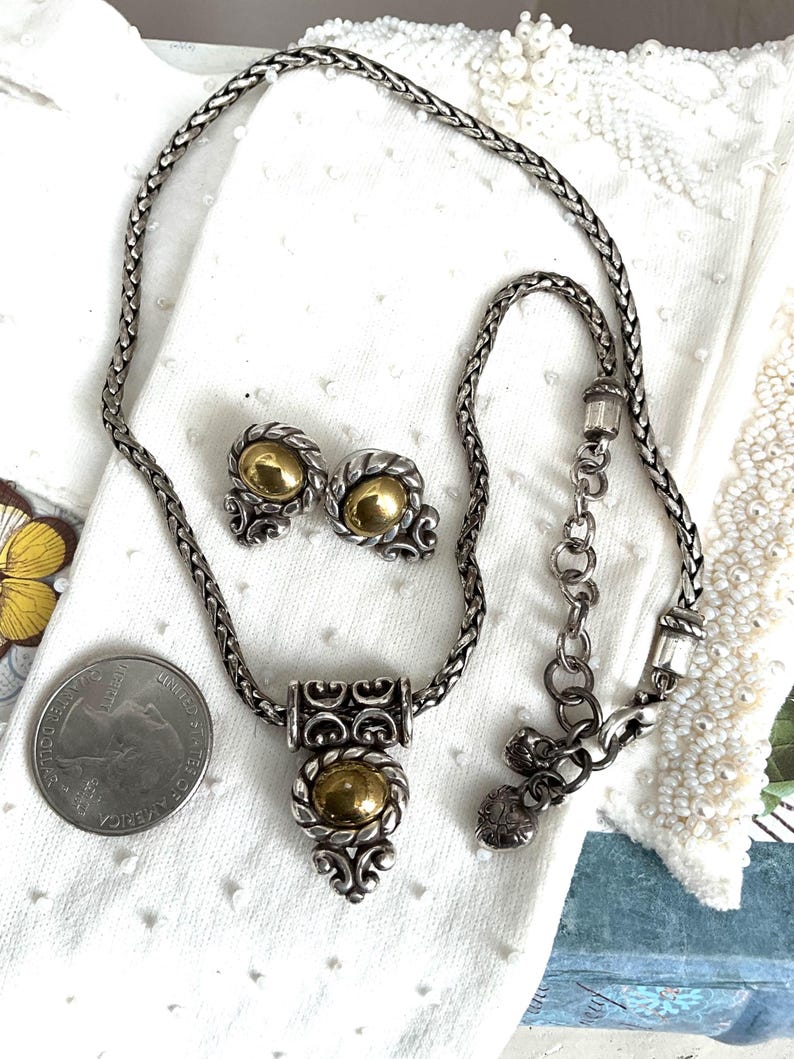 May include: A silver-toned necklace and matching earrings set with gold-colored accents. The necklace features a braided chain and a pendant with an oval gold center. The earrings have a similar design. A US quarter is next to the jewelry.