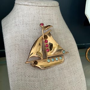 May include: A gold sailboat brooch with pink and blue rhinestones. The sailboat has a white sail and a red flag.