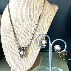 May include: A silver-toned necklace and earring set. The necklace features a pendant with a central pearl and ornate detailing. The earrings, displayed on a stand, mirror the pendant design. The jewelry is displayed on a neutral-toned bust.