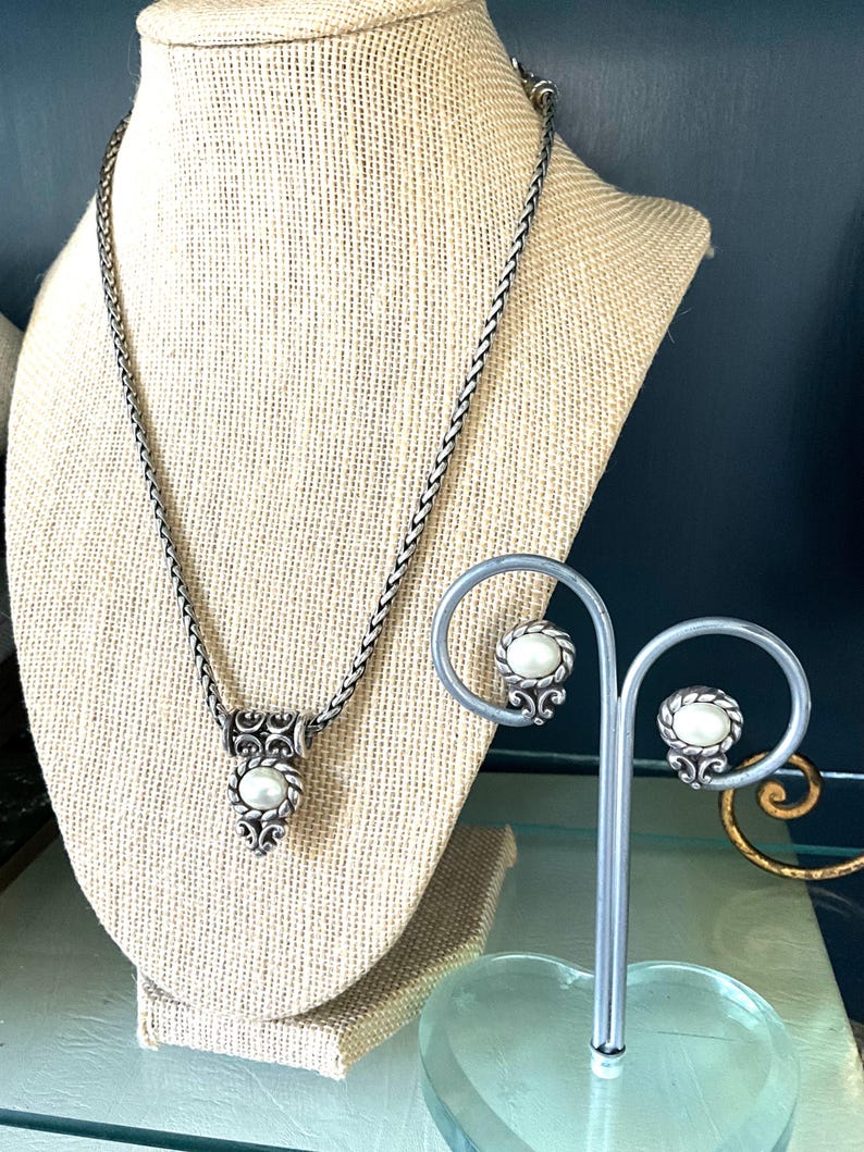 May include: A silver-toned necklace and earring set. The necklace features a pendant with a central pearl and ornate detailing. The earrings, displayed on a stand, mirror the pendant design. The jewelry is displayed on a neutral-toned bust.