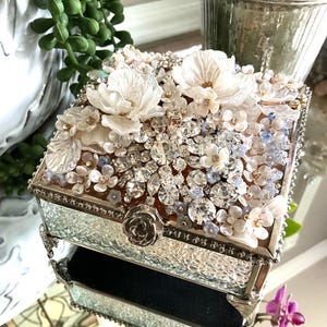 May include: A decorative, square jewelry box with a mirrored, textured glass exterior. The top is adorned with white floral embellishments, clear rhinestones, and small blue accents. A silver rose detail is on the front. The box is open, revealing a black velvet interior.