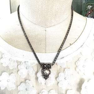 May include: A silver-toned necklace with a braided chain and a pendant featuring a pearl. The pendant has an ornate design with scrollwork and a central pearl. The necklace is displayed on a mannequin.