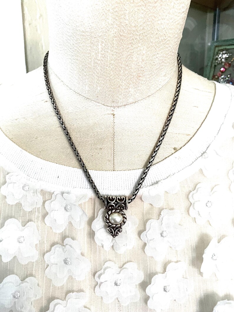 May include: A silver-toned necklace with a braided chain and a pendant featuring a pearl. The pendant has an ornate design with scrollwork and a central pearl. The necklace is displayed on a mannequin.
