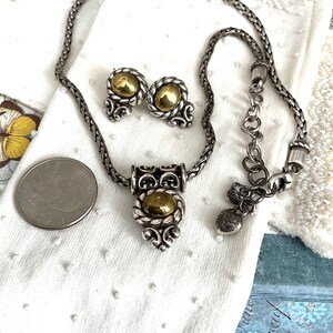 May include: A silver-toned necklace and matching earrings set with gold-colored accents. The necklace features a pendant with a central gold-colored stone and decorative silver filigree. The earrings have a similar design. The set is displayed on a white surface.