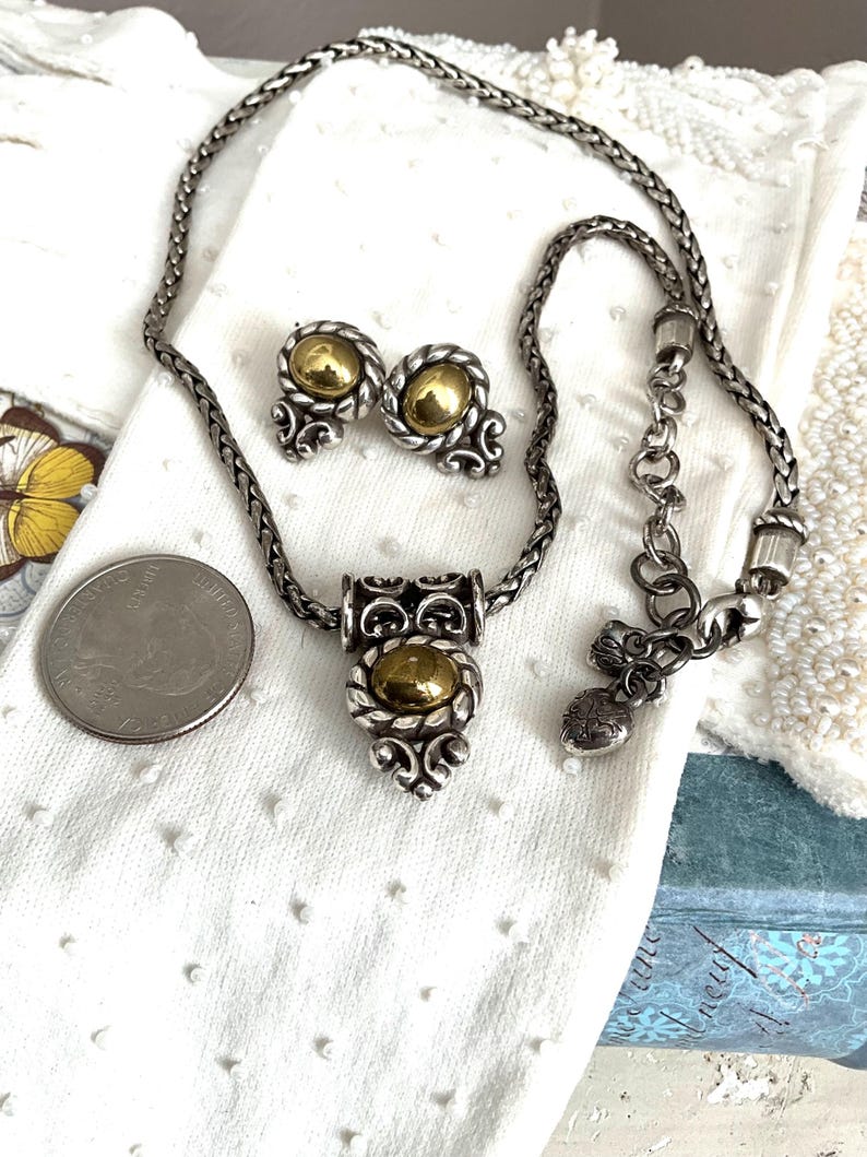 May include: A silver-toned necklace and matching earrings set with gold-colored accents. The necklace features a pendant with a central gold-colored stone and decorative silver filigree. The earrings have a similar design. The set is displayed on a white surface.