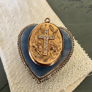 May include: A gold locket with a cross design set with clear rhinestones. The locket is in a blue velvet heart-shaped box.
