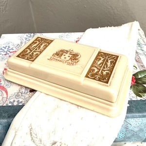 May include: A rectangular, off-white box with gold-colored decorative accents and the name "HAMILTON" printed on the lid. The box features ornate designs and a crest-like emblem. The box is likely vintage and sits on a patterned surface.