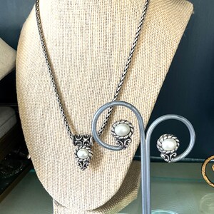May include: A silver-toned necklace and matching earrings set. The necklace features a pendant with a central pearl and ornate detailing. The earrings have a similar design. The jewelry is displayed on a beige mannequin.