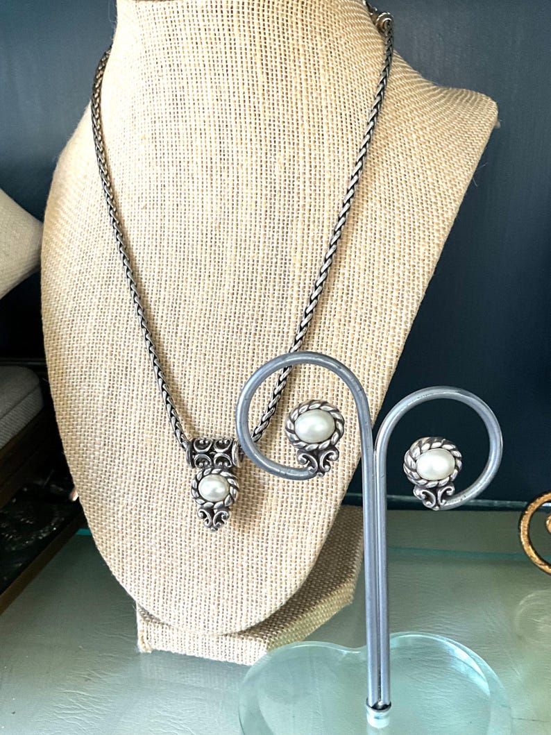 May include: A silver-toned necklace and matching earrings set. The necklace features a pendant with a central pearl and ornate detailing. The earrings have a similar design. The jewelry is displayed on a beige mannequin.