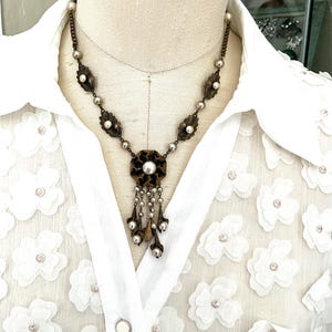 May include: A necklace featuring a bronze-toned chain with alternating metal leaf motifs and small round white beads. The central pendant is a circular bronze flower element with a large white bead, accented by dangling chains with beads.