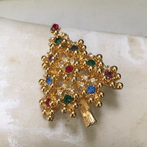 May include: A gold-toned Christmas tree brooch with a textured surface. The brooch is adorned with colorful gemstones in red, green, blue, and purple, and clear rhinestones. The tree trunk is a solid gold color.