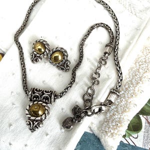 May include: A silver-toned necklace and matching earrings set. The necklace features a braided chain with a pendant and a gold-colored orb. The earrings have a similar design with gold-colored orbs. The jewelry set is displayed on a white surface.