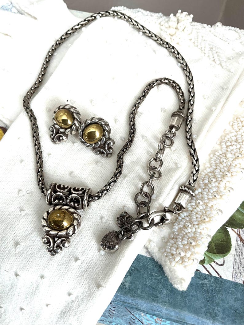 May include: A silver-toned necklace and matching earrings set. The necklace features a braided chain with a pendant and a gold-colored orb. The earrings have a similar design with gold-colored orbs. The jewelry set is displayed on a white surface.