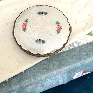May include: A round silver compact case with a scalloped edge and a guilloche pattern. The top features a central embossed design surrounded by four hand-painted floral motifs in pink, blue, and green enamel. The metal sides have a dark patina.