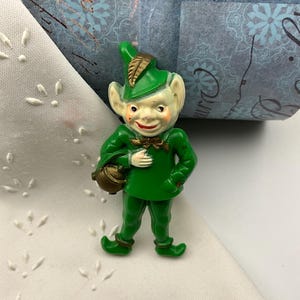 May include: A vintage figurine of a green elf with a gold leaf on its hat and a small brown pot. The elf has a smiling face with rosy cheeks and is wearing a green outfit. The figurine is on a white cloth with a floral pattern.