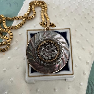 May include: A gold chain necklace with a large, round pendant. The pendant is silver and has a raised design of a flower with rhinestones around it. The pendant is in a small, white box with a blue velvet lining.