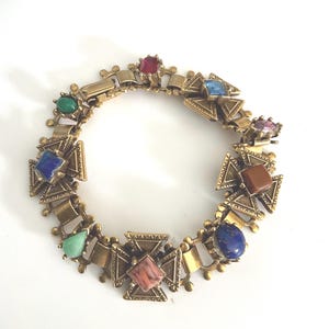 May include: A vintage gold-tone bracelet featuring square and triangular decorative elements. The bracelet is adorned with various colorful gemstones, including red, blue, green, and purple. The bracelet is displayed on a white surface.
