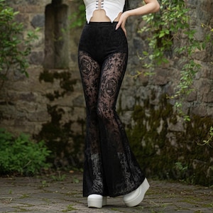 May include: A white halter top with a lace-up front and black lace bell-bottom pants. The pants have a floral pattern. The model is wearing white platform shoes. The background is a stone wall with greenery.
