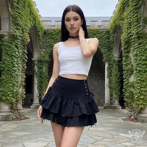 May include: A woman wearing a white tank top and a black ruffled mini skirt with lace-up detailing. The skirt has two tiers of ruffles. The background features a stone structure covered in green vines.