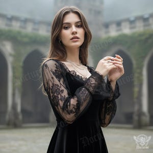 May include: A black velvet dress with long sleeves featuring intricate black lace detailing. The dress has a scoop neckline and a flowing skirt. The sleeves are sheer, showcasing the lace pattern. The background is a blurred outdoor setting with a stone building.