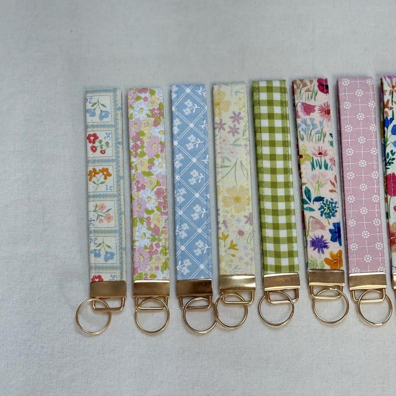 May include: A collection of key fobs with various floral and patterned designs. Each fob features a gold-colored metal clasp and ring. The fabric patterns include flowers, gingham, and geometric shapes in pastel colors.