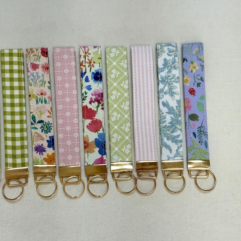 May include: A collection of eight fabric key fobs with gold-colored hardware. The key fobs feature various floral and patterned designs in shades of green, pink, blue, and purple. Each fob is approximately 6 inches long.