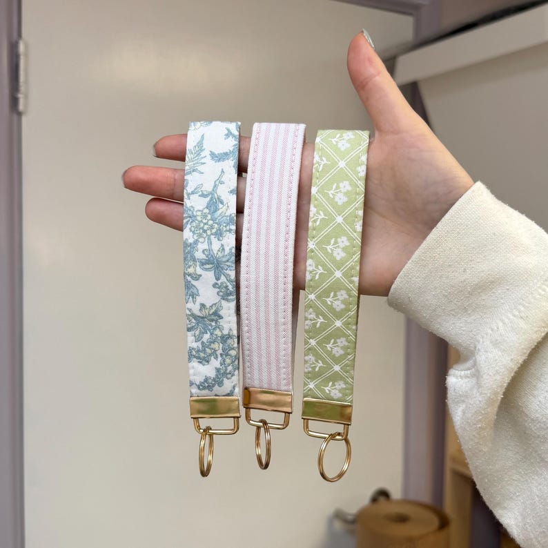 May include: Three fabric wrist keychains in the image. One is white with blue floral print, the second is pink with vertical stripes, and the third is green with a white floral pattern. Each keychain has a gold-colored metal clasp and ring.