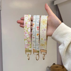 May include: Three fabric key fobs with floral patterns in pink, blue, and yellow. Each fob has a gold-colored metal clasp and ring. The key fobs are held up against a neutral background.