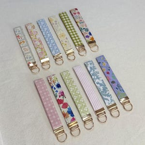 May include: A collection of fabric key fobs in various floral and patterned designs. Each fob features a gold-colored metal clasp and ring. The color palette includes pink, blue, green, and purple, with a white background.