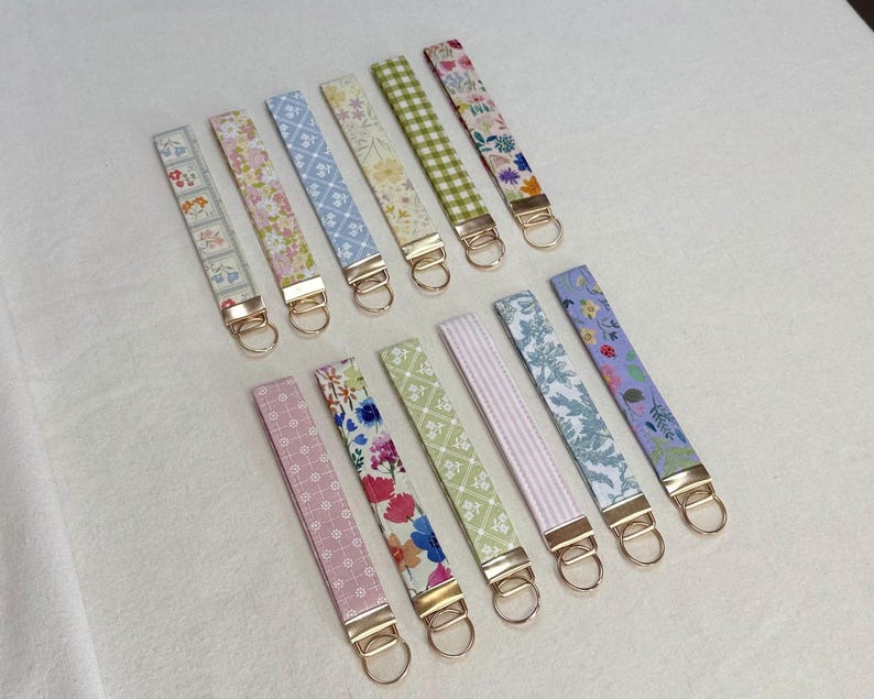 May include: A collection of fabric key fobs in various floral and patterned designs. Each fob features a gold-colored metal clasp and ring. The color palette includes pink, blue, green, and purple, with a white background.