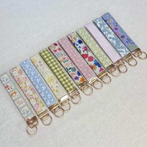 May include: A collection of fabric key fobs with various floral and patterned designs. Each fob features a gold-colored metal clasp and key ring. The fabrics display a range of colors, including pink, blue, green, and white, with floral, striped, and checkered patterns.