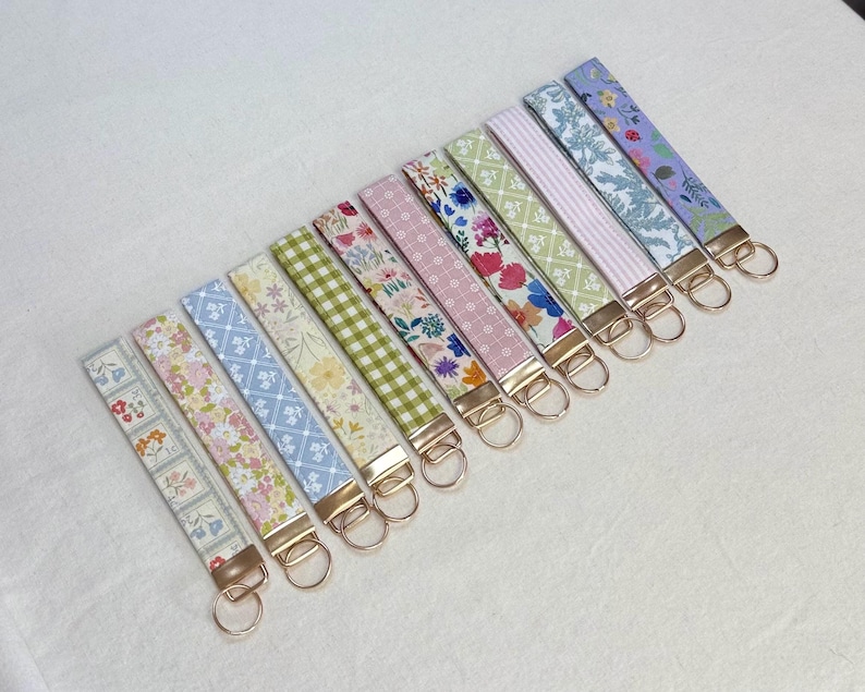 May include: A collection of fabric key fobs with various floral and patterned designs. Each fob features a gold-colored metal clasp and key ring. The fabrics display a range of colors, including pink, blue, green, and white, with floral, striped, and checkered patterns.