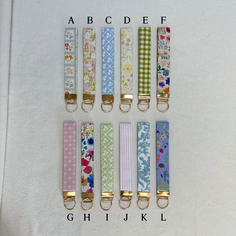 May include: A collection of twelve fabric wrist keychains in various floral and patterned designs. Each keychain features a gold-colored metal ring and clasp. The patterns include flowers, gingham, stripes, and geometric shapes in colors like pink, blue, green, and purple.