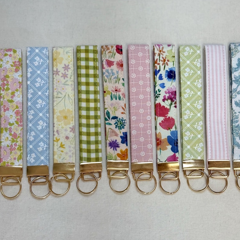 May include: A collection of fabric key fobs with various floral and patterned designs. Each fob features a gold-colored metal ring and a matching gold-colored metal rectangle at the top. The patterns include floral prints, gingham, and geometric designs.