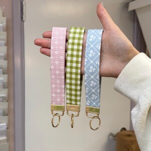 May include: Three fabric keychains in pink floral, green gingham, and blue floral patterns. Each keychain has a gold-colored metal clasp and ring. The keychains are held up in a hand.